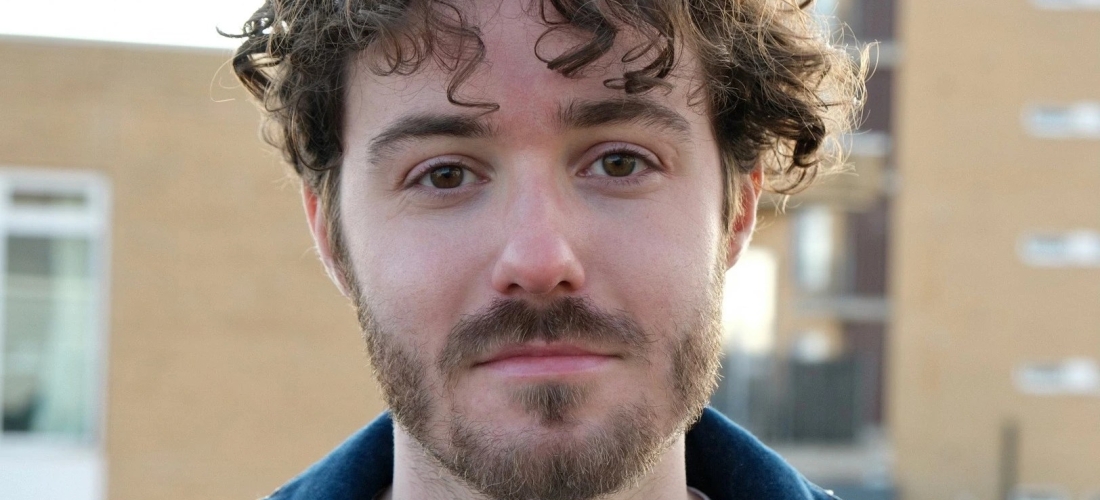 a photo of sam barker slightly smirking, with his curly hair framed by the light from behind